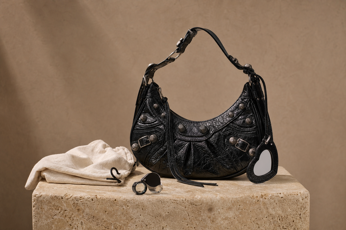 Editorial still life of an unbranded black crinkled-leather hobo bag with silver-tone studs, dust pouch, and inspection loupe for a pre-owned Balenciaga Le Cagole buying guide.
