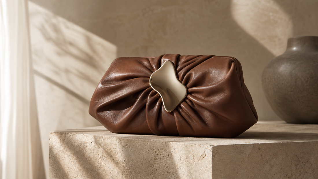 Editorial still life of an unbranded soft brown leather clutch with an abstract metal clasp on a warm stone pedestal