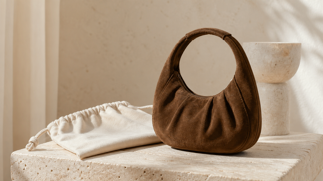 Unbranded brown suede hobo bag with a rounded top handle on a warm stone surface beside a plain dust bag.